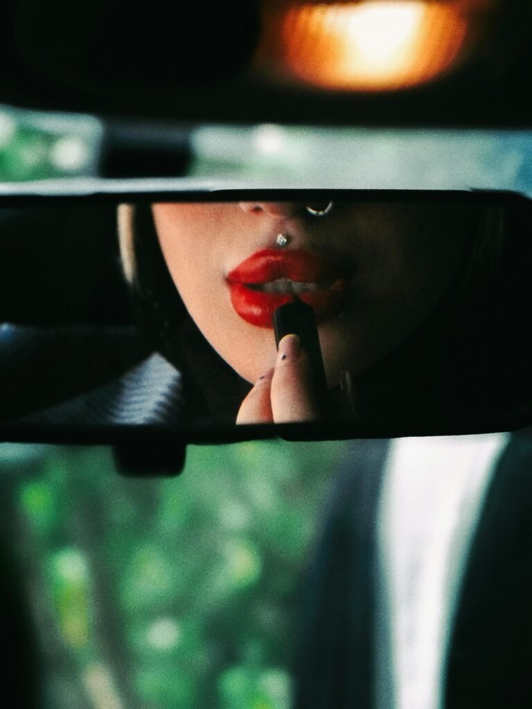 A stylish woman applies bold red lipstick, reflected in a car's rearview mirror.