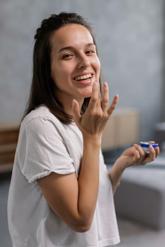 Photo by Alena Darmel A cheerful woman applying moisturizing cream with a blurred background, indoors.